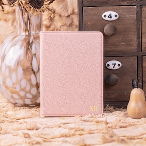 以下が含まれることがあります： 右下に金色のイニシャル「SB」が入った、明るいピンク色の革風ジャーナル。ジャーナルは、乾燥したヒマワリの花が入った花瓶と木製の梨の置物を背景に、テクスチャ加工された表面に立ててあります。