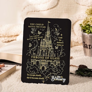 Könnte beinhalten: Eine schwarze, rechteckige E-Reader-Hülle mit goldenen Linienzeichnungen eines Schlosses, Vögeln und Schmetterlingen. Der Text lautet: YOU COULD RATTLE THE STARS, Until the darkness claims us, THRONE OF GLASS, To escape death, she’d become death und I do not yield.