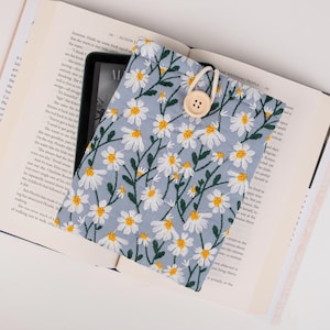 May include: A light blue fabric pouch with a white daisy floral pattern. The pouch has a drawstring closure and a wooden button. The pouch is lying on top of an open book.