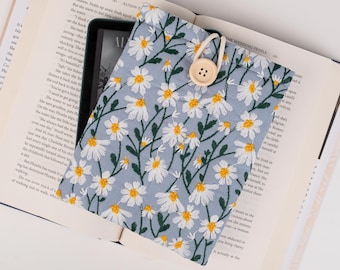 May include: A light blue fabric pouch with a white daisy floral pattern. The pouch has a drawstring closure and a wooden button. The pouch is lying on top of an open book.