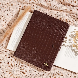 May include: A brown leather journal with a crocodile texture, featuring a gold-colored initial monogram "SB". The journal is closed with a snap closure and rests on an open book. The background is a cream-colored, crocheted textile.