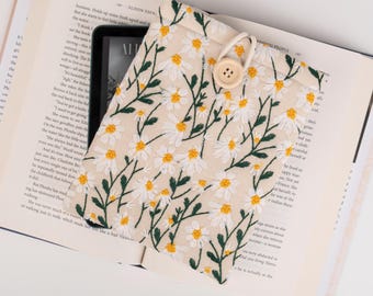 Pode incluir: Uma capa para leitor de e-books em tecido bege com um padrão floral de margaridas brancas. A capa tem um botão castanho e um fecho de cordão.