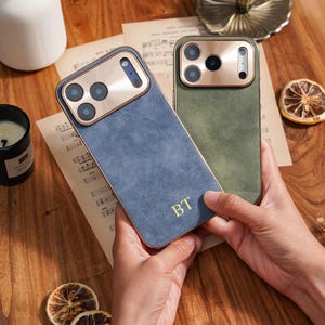 Op de afbeelding: Twee telefoonhoesjes, &eacute;&eacute;n blauw en &eacute;&eacute;n groen, met goudkleurige camera-omrandingen en randen. De blauwe hoes heeft de initialen "BT" in goud. Ze liggen op een houten oppervlak met gedroogde citrusvruchten.