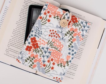 Op de afbeelding: Een witte e-reader hoes van stof met een geborduurd bloemenmotief. De borduursels bevatten roze, rode en blauwe bloemen en bladeren op een witte achtergrond. Een houten knoop is aan de hoes bevestigd.