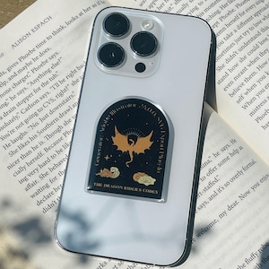 以下が含まれることがあります： 金色のドラゴン、雲、そして「THE DRAGON RIDER'S CODEX」の文字が描かれたステッカーが付いた透明な携帯電話ケース。 電話は、テキストが書かれた開いた本の上に置かれています。