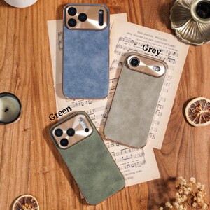 Op de afbeelding: Drie telefoonhoesjes in blauw, grijs en groen, met goudkleurige camerabehuizingen, worden getoond op een houten oppervlak. De hoesjes liggen op vintage bladmuziek. De woorden "Blue", "Grey" en "Green" staan boven elke hoes.