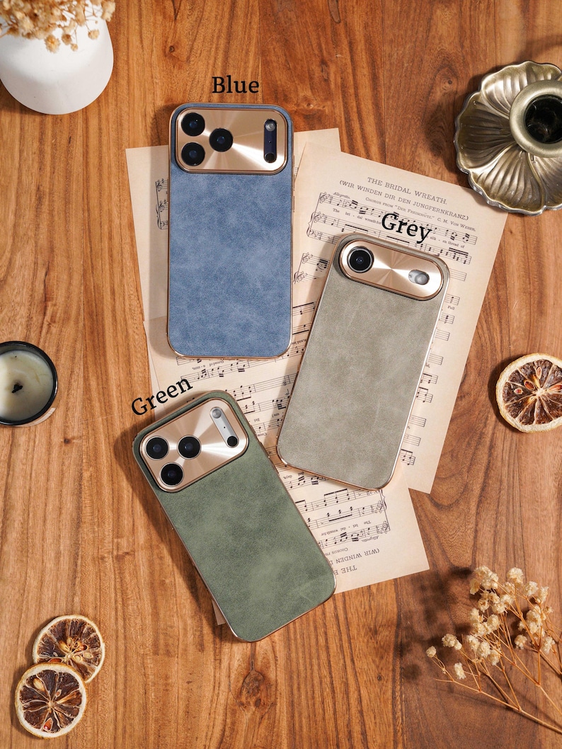 Op de afbeelding: Drie telefoonhoesjes in blauw, grijs en groen, met goudkleurige camerabehuizingen, worden getoond op een houten oppervlak. De hoesjes liggen op vintage bladmuziek. De woorden "Blue", "Grey" en "Green" staan boven elke hoes.
