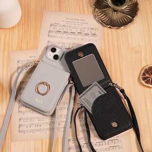 Könnte beinhalten: Zwei Handyhüllen mit Umhängeriemen. Eine ist hellgrau mit einem goldenen Ring und den Initialen "KO". Die andere ist schwarz mit einem Spiegel und einer Karte. Beide Hüllen liegen auf Notenblättern.