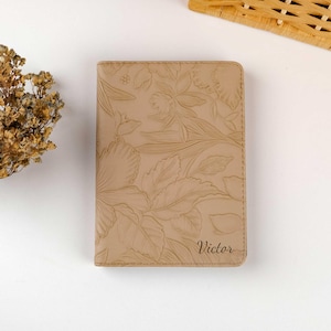 May include: A beige leather passport holder with a floral embossed design and the name "Victor" engraved on the front.