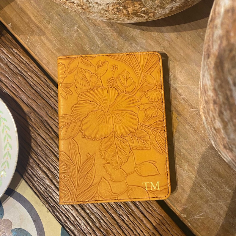 May include: A tan leather passport holder with an embossed floral design and the initials "TM" on the bottom right corner.