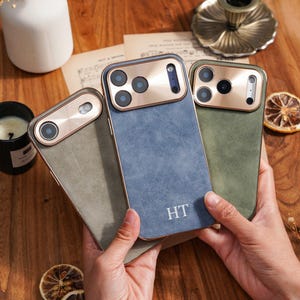 Op de afbeelding: Drie telefoonhoesjes in groen en blauw, met een goudkleurig camerakader. De hoesjes hebben een getextureerde afwerking en de initialen "HT" zijn zichtbaar op het blauwe hoesje. De hoesjes worden in iemands handen gehouden.