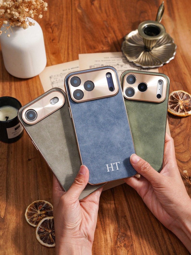 Op de afbeelding: Drie telefoonhoesjes in groen en blauw, met een goudkleurig camerakader. De hoesjes hebben een getextureerde afwerking en de initialen "HT" zijn zichtbaar op het blauwe hoesje. De hoesjes worden in iemands handen gehouden.