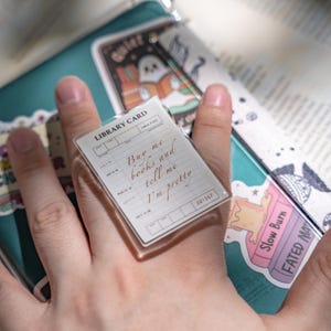 Può includere: Un anello in acrilico trasparente con un design di scheda di biblioteca. La scheda riporta la scritta "Buy me books and tell me I'm pretty" in corsivo marrone. L'anello è tenuto sopra un taccuino turchese con adesivi.