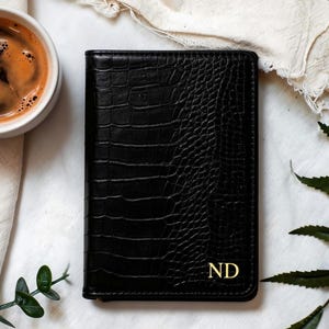 May include: Black leather wallet with a crocodile skin texture, featuring gold-colored initials "ND". The wallet is rectangular and closed, with a smooth, shiny finish. A cup of coffee sits to the left of the wallet.