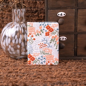 May include: A rectangular notebook cover featuring a white fabric base with intricate embroidery in shades of orange, peach, and light blue. The floral and botanical patterns cover the entire surface, showcasing detailed stitching and textures.
