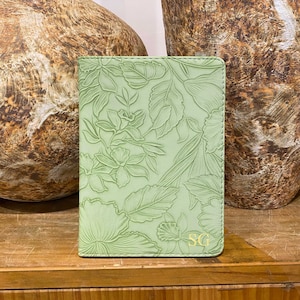 May include: A light green leather passport holder with a floral embossed design and the initials "SG" in gold lettering.