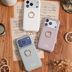 Op de afbeelding: Drie gepersonaliseerde telefoonhoesjes in wit, lavendel en grijs, elk met een gouden ringhouder en initialen. De hoesjes liggen op een houten oppervlak met bladmuziek en gedroogde citrusvruchten.