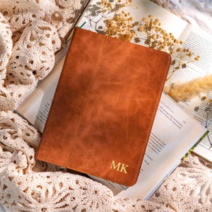 Puede incluir: Una funda de tableta de cuero marrón con las iniciales doradas "MK". La funda está sobre un libro abierto con flores secas. La funda tiene una costura cosida en el medio. La funda mide unos 25 cm de largo.