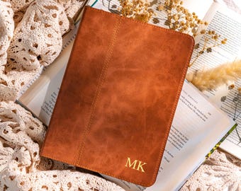 May include: A brown leather tablet case with the initials "MK" in gold lettering. The case is lying on top of an open book with dried flowers. The case has a stitched seam down the middle.