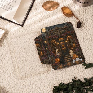 May include: A clear plastic e-reader case featuring a detailed illustration of a storefront titled The Raven & Quill Bookshop, with ghosts, pumpkins, a black cat, and the name Brittany printed in white script on the lower right corner.