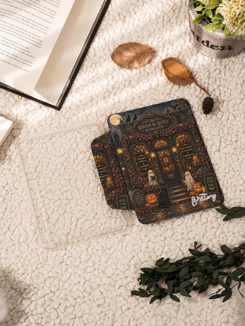 May include: A clear plastic e-reader case featuring a detailed illustration of a storefront titled The Raven & Quill Bookshop, with ghosts, pumpkins, a black cat, and the name Brittany printed in white script on the lower right corner.