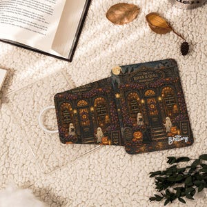 May include: Two rectangular e-reader cases featuring a detailed illustration of The Raven & Quill Bookshop with ghosts, pumpkins, and a black cat. The design includes text reading The Raven & Quill Bookshop, Books Spells & Stories, and Read If You Dare.