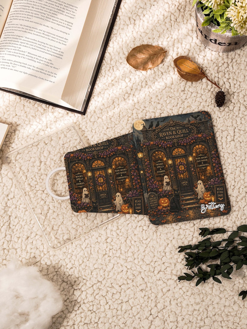May include: Two rectangular e-reader cases featuring a detailed illustration of The Raven & Quill Bookshop with ghosts, pumpkins, and a black cat. The design includes text reading The Raven & Quill Bookshop, Books Spells & Stories, and Read If You Dare.
