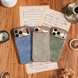 Op de afbeelding: Drie telefoonhoesjes in blauw, grijs en groen met goudkleurige camerakaders liggen op een houten oppervlak met bladmuziek. Gedroogde bloemen, een kaars en decoratieve elementen completeren de sc&egrave;ne. De tekst op de bladmuziek luidt "BUY A DROOM, Concluded" en "THE BRIDAL WREATH."