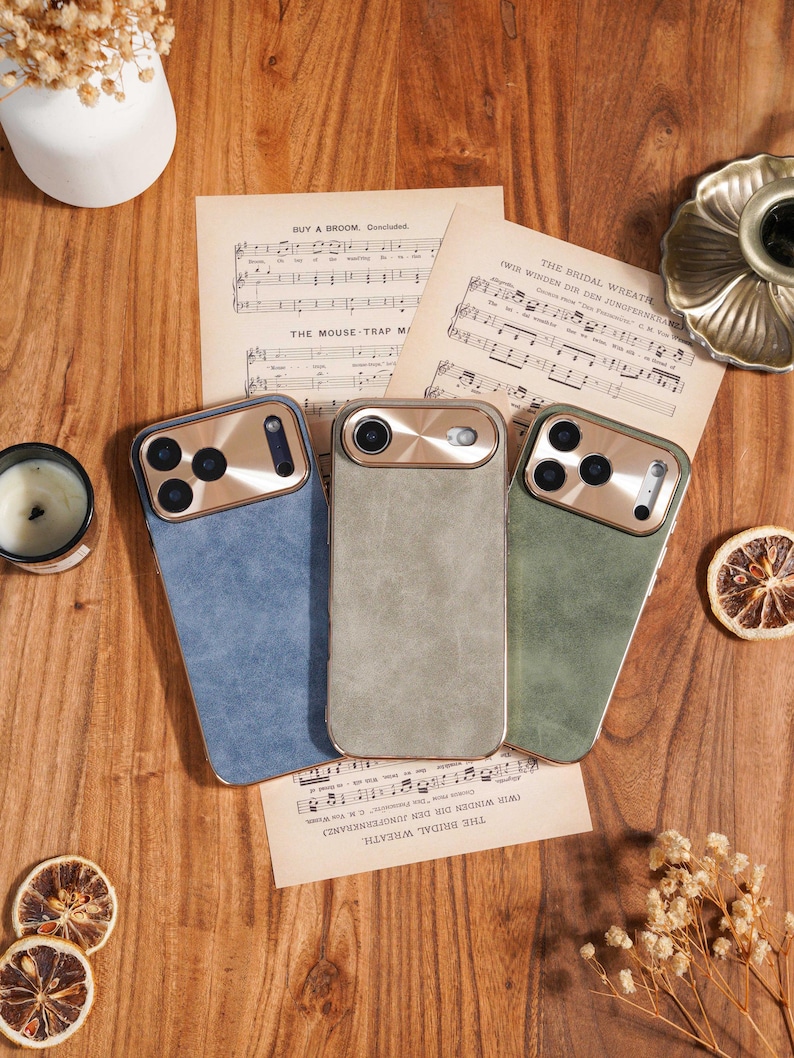 Op de afbeelding: Drie telefoonhoesjes in blauw, grijs en groen met goudkleurige camerakaders liggen op een houten oppervlak met bladmuziek. Gedroogde bloemen, een kaars en decoratieve elementen completeren de sc&egrave;ne. De tekst op de bladmuziek luidt "BUY A DROOM, Concluded" en "THE BRIDAL WREATH."