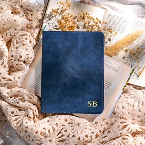 May include: A navy blue leather journal with the gold initials "SB" is displayed open on a table. The journal is surrounded by open books, dried flowers, and a cream-colored lace cloth. The journal has a stitched spine.