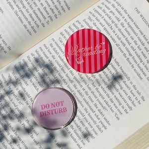 Pu&ograve; includere: Due bottoni rotondi, uno rosa e bianco a righe con il testo "Rather be reading" e un disegno di libro, l'altro rosa con il testo "DO NOT DISTURB". Entrambi sono su una pagina di libro.