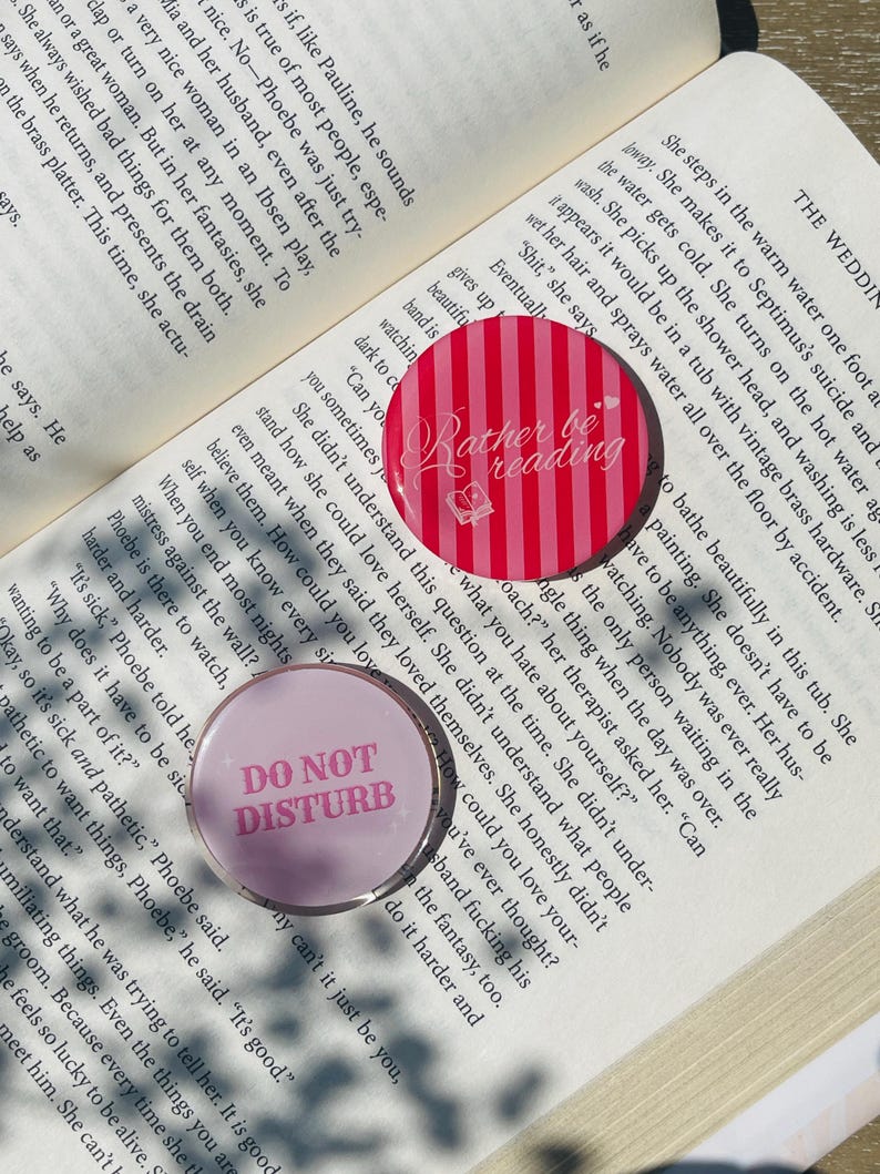 Pu&ograve; includere: Due bottoni rotondi, uno rosa e bianco a righe con il testo "Rather be reading" e un disegno di libro, l'altro rosa con il testo "DO NOT DISTURB". Entrambi sono su una pagina di libro.