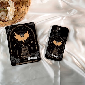 Może przedstawiać: Czarne etui na telefon i tablet z złotym motywem smoka nad stos książek. Tekst zawiera "Brittney" i "The Dragon Rider's Codex". Projekt jest na czarnym tle z niebiańskimi akcentami.