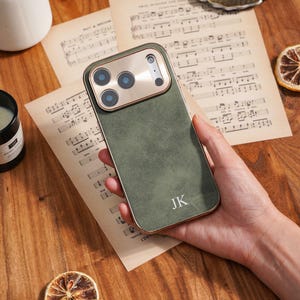 May include: A green phone case with a gold-coloured camera module and trim. The case has the initials "JK" printed on the lower right corner. The phone case is resting on a wooden surface.