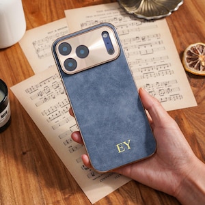 Op de afbeelding: Een blauwe telefoonhoes met een gouden cameramodule en rand. De hoes heeft de initialen "EY" in gouden letters. De telefoonhoes wordt in een hand gehouden en ligt op bladmuziek.