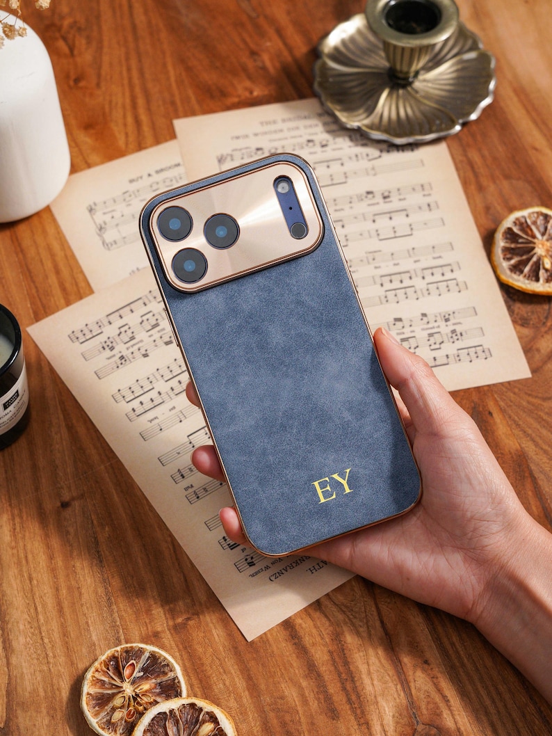Op de afbeelding: Een blauwe telefoonhoes met een gouden cameramodule en rand. De hoes heeft de initialen "EY" in gouden letters. De telefoonhoes wordt in een hand gehouden en ligt op bladmuziek.