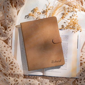May include: A tan leather journal with a snap closure, resting on an open book. The journal is embossed with the name "Richard" in a cursive font. The background includes a lace cloth and dried flowers.