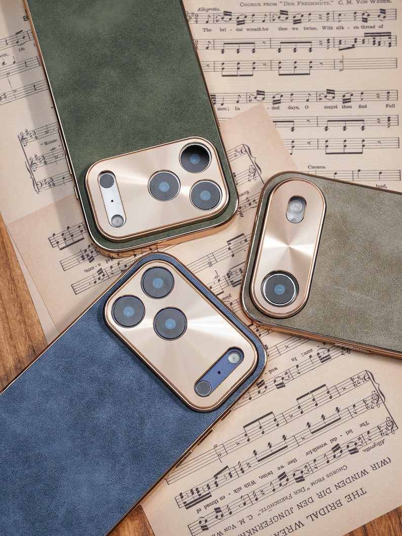 Op de afbeelding: Close-up van verschillende telefoonhoesjes in groen en blauw, met goudkleurige camerakaders. De hoesjes zijn gerangschikt op een achtergrond van vintage bladmuziek, wat een klassieke esthetiek laat zien. De hoesjes hebben een getextureerde uitstraling.