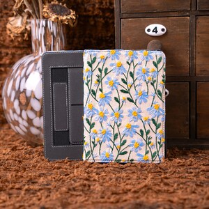 Peut inclure: Un étui de tablette gris avec un intérieur noir et une couverture crème ornée de broderies de fleurs de marguerites bleues et jaunes. L'étui est ouvert, révélant le support de la tablette.