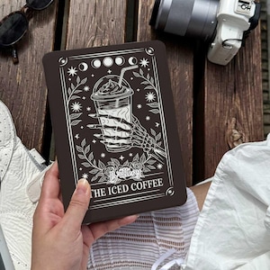 May include: A dark brown tablet case with a white illustration of a skeleton hand holding an iced coffee. The design includes stars, moons, and floral elements. The text "Brittany" and "THE ICED COFFEE" are also visible.