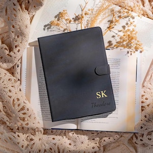 Può includere: Un diario in pelle nera con chiusura dorata, appoggiato su un libro aperto. Il diario è personalizzato con le iniziali "SK" e il nome "Theodore" in lettere dorate. Fiori secchi e un panno di pizzo sullo sfondo.