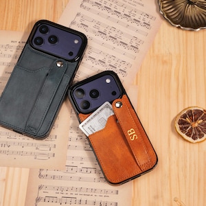 Puede incluir: Dos fundas de teléfono con tarjeteros. Una es verde azulado, la otra es marrón con las iniciales "SB". Ambas fundas tienen un marco negro y una tarjeta insertada en el tarjetero. Las fundas están sobre una superficie de madera con partituras.
