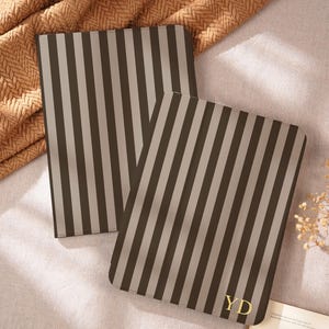 May include: Two rectangular tablet cases featuring a vertical striped pattern in dark brown and light beige. The front case displays the gold-colored initials YD in the bottom right corner. Both cases have rounded corners and a smooth, flat surface.