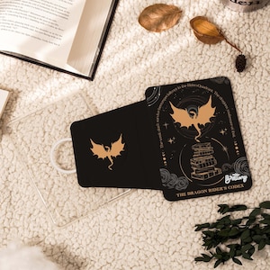Puede incluir: Tarjeta negra con una ilustración de dragón dorado y el texto "THE DRAGON RIDER'S CODEX". La tarjeta está abierta, revelando una pila de libros y diseños celestiales. También se ve una funda transparente para teléfono y un libro.