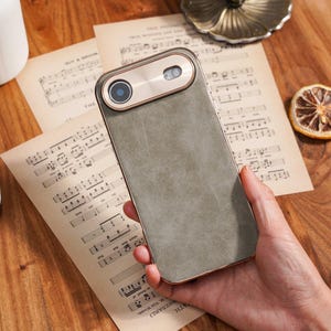 Op de afbeelding: Een smartphonehoesje met een grijze leerachtige afwerking en een goudkleurige cameramodule. De hoes heeft een rechthoekige vorm met afgeronde hoeken. De cameramodule heeft een ronde lens en een goudkleurige rand. De hoes ligt op bladmuziek.