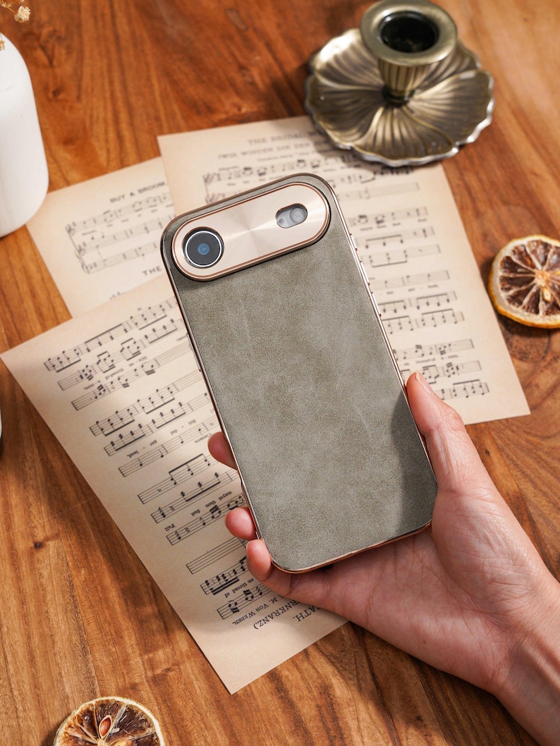Op de afbeelding: Een smartphonehoesje met een grijze leerachtige afwerking en een goudkleurige cameramodule. De hoes heeft een rechthoekige vorm met afgeronde hoeken. De cameramodule heeft een ronde lens en een goudkleurige rand. De hoes ligt op bladmuziek.