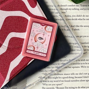 May include: A pink tarot card featuring a crescent moon with an eye, clouds, and stars. The card is labeled "The Moon" and has a pink border. The card is in a clear plastic case.