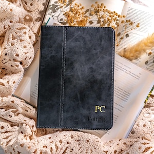 May include: A dark gray leather tablet case with the initials "PC" and the name "Patrick" in gold lettering. The case is lying on an open book and a cream-colored crocheted cloth.