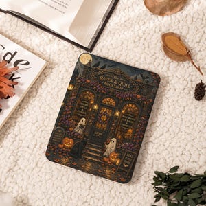 May include: A rectangular e-reader cover featuring a detailed illustration of a bookshop named The Raven & Quill Bookshop. The scene includes a black cat, two ghosts, pumpkins, autumn leaves, and text reading Books Spells & Stories and Read If You Dare.