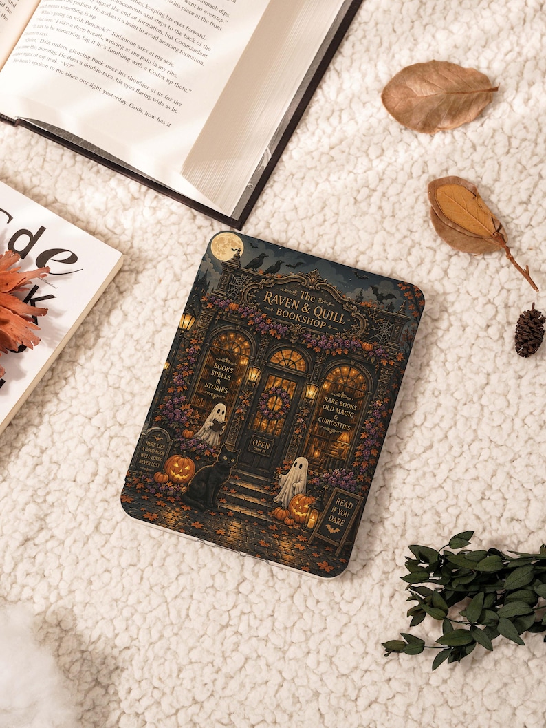 May include: A rectangular e-reader cover featuring a detailed illustration of a bookshop named The Raven & Quill Bookshop. The scene includes a black cat, two ghosts, pumpkins, autumn leaves, and text reading Books Spells & Stories and Read If You Dare.